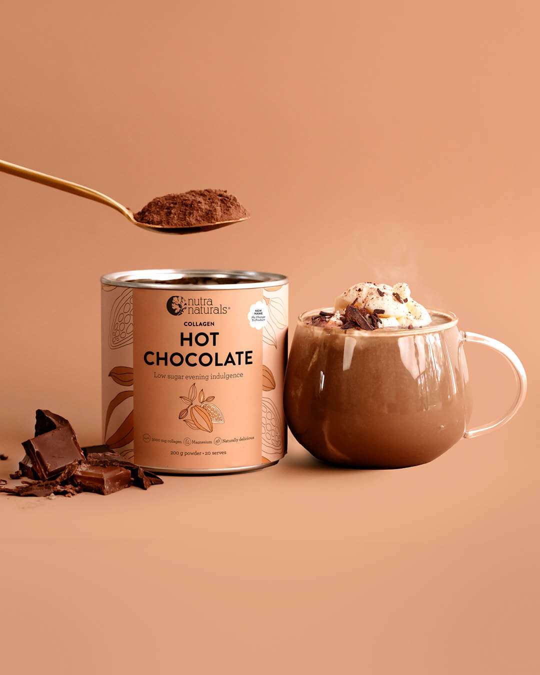 A can of Collagen Hot Chocolate powder stands next to a mug filled with hot chocolate topped with whipped cream and chocolate shavings, with a gold spoon holding cocoa powder above the can on a warm beige background.