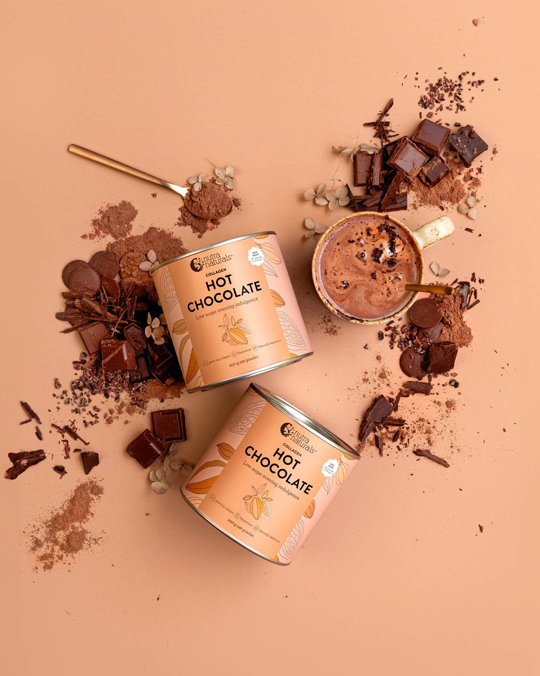 Two cans of Collagen Hot Chocolate are arranged on a peach background with chocolate pieces, cocoa powder, and a mug of hot chocolate.