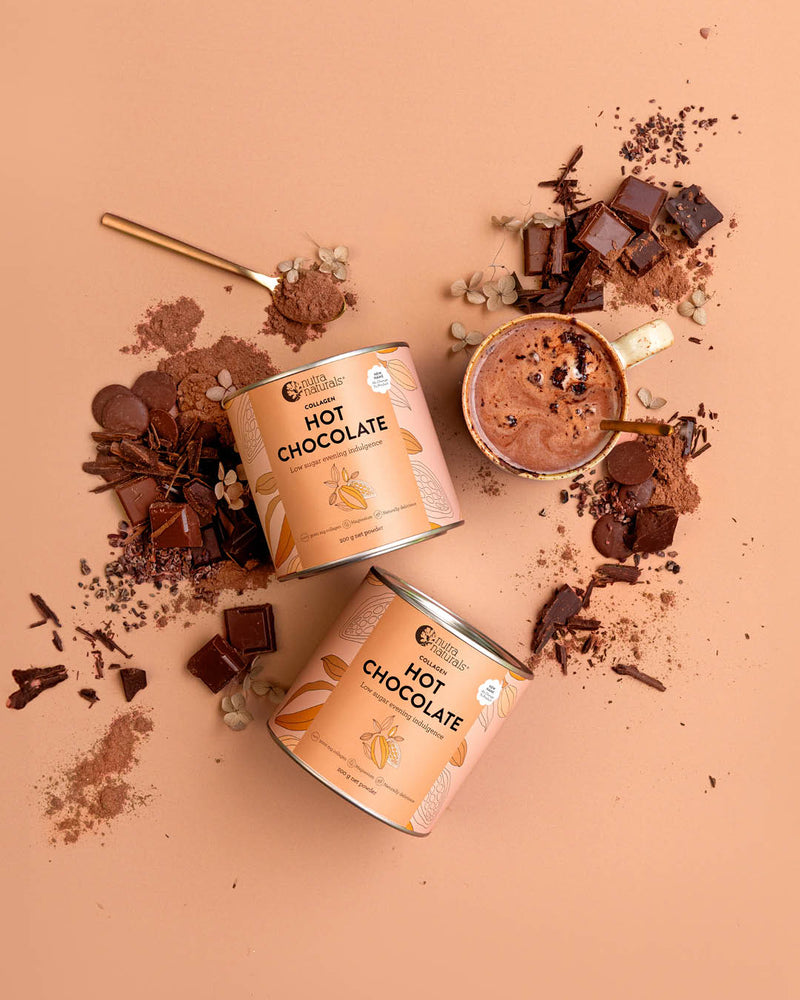Two cans of Collagen Hot Chocolate are arranged on a peach background with chocolate pieces, cocoa powder, and a mug of hot chocolate.