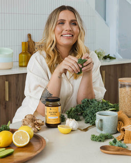 A woman is smiling with a jar of beef bone broth lemon ginger ACV in front of her