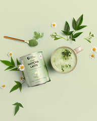 A can of Collagen Matcha Latte powder from is styled in a flat-lay composition with a prepared matcha latte in a beige mug, a gold spoon with green powder, and fresh green leaves and daisies on a pale sage green background.