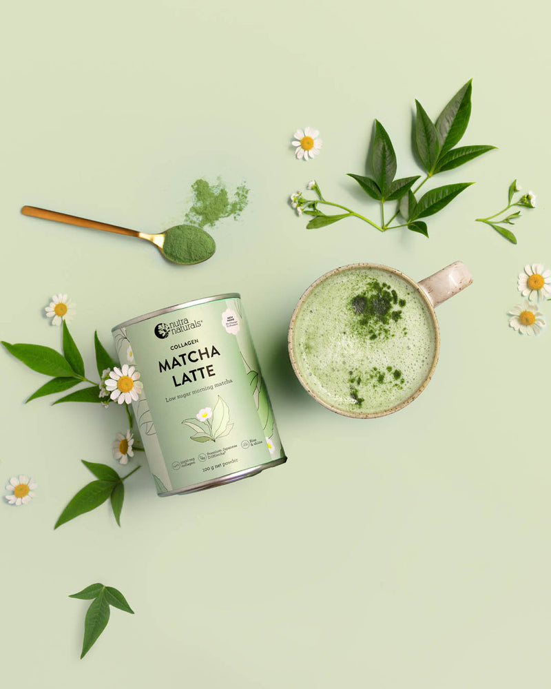 A can of Collagen Matcha Latte powder from is styled in a flat-lay composition with a prepared matcha latte in a beige mug, a gold spoon with green powder, and fresh green leaves and daisies on a pale sage green background.