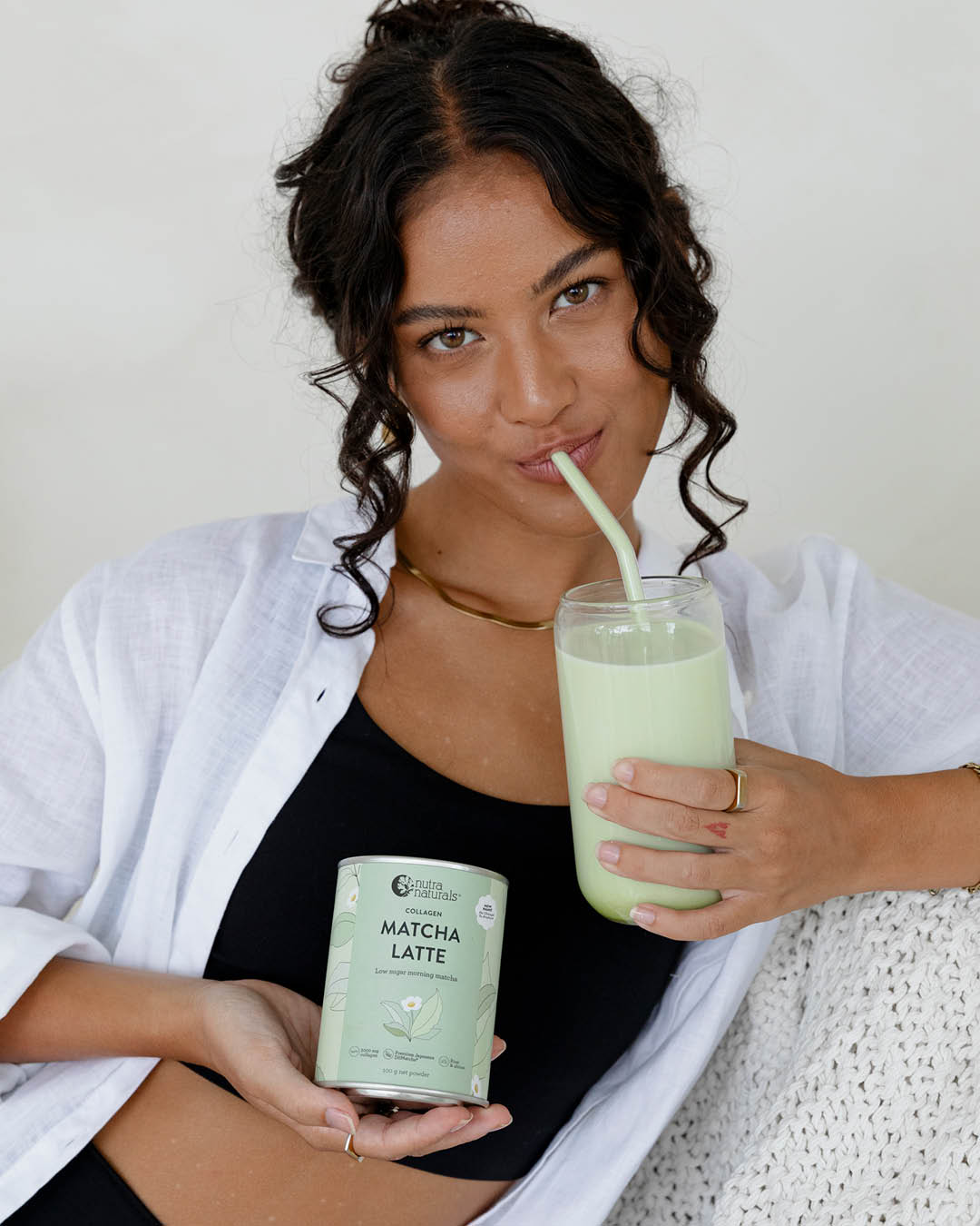 A woman holding a glass of matcha latte with a straw and a can of Collagen Matcha Latte powder