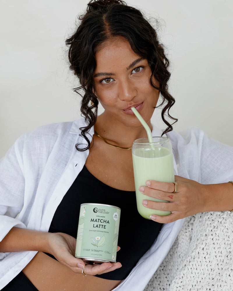 A woman holding a glass of matcha latte with a straw and a can of Collagen Matcha Latte powder