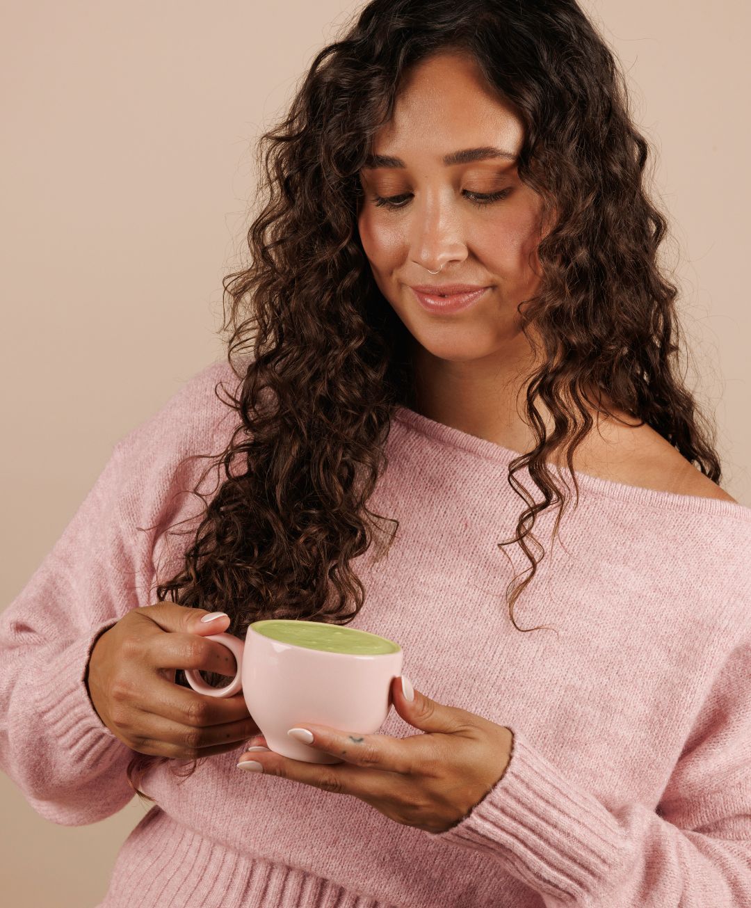 Woman in a pink sweater holding a white mug with a green beverage against a beige background