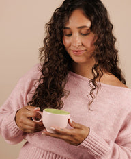 Woman in a pink sweater holding a white mug with a green beverage against a beige background