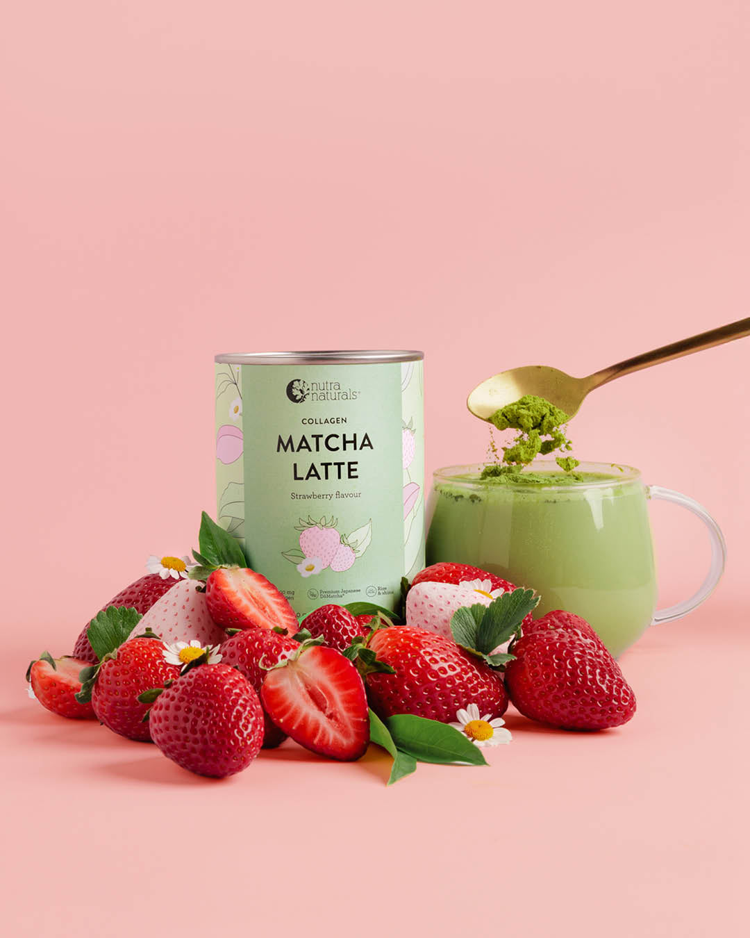 A can of Collagen Matcha Latte Strawberry flavor stands next to a glass mug filled with green matcha and topped with a gold spoonful, surrounded by fresh strawberries on a solid pink background.