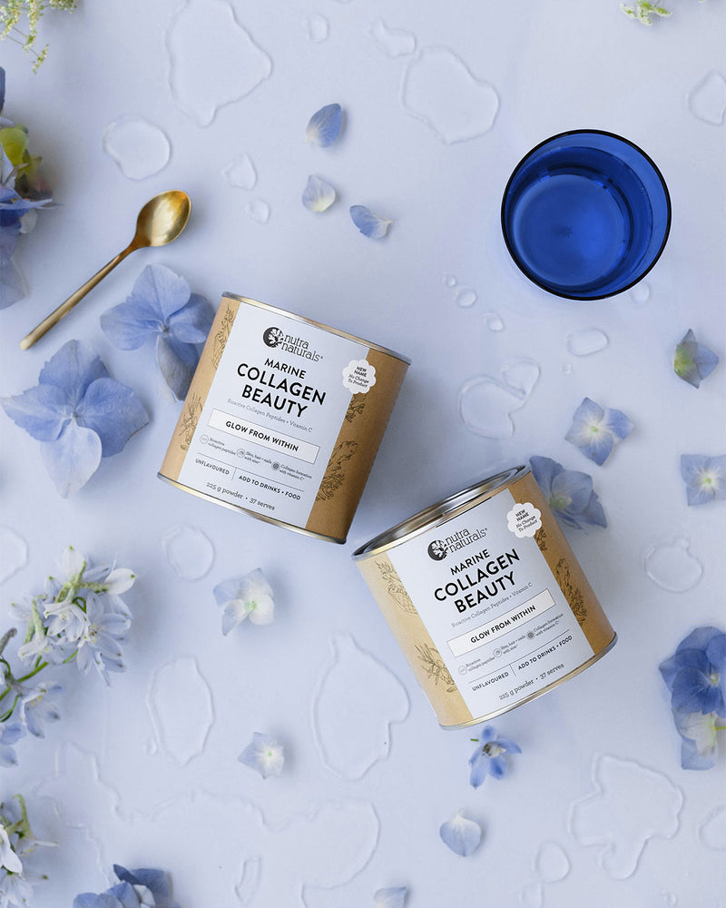 Two cans of Collagen Beauty powder arranged on a pale blue surface with scattered blue flower petals, a gold spoon, and a blue glass.
