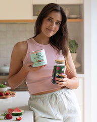 A woman is holding a can of Super Greens + Reds Watermelon Strawberry powder and a dark green glass bottle with Super Greens + Reds Watermelon Strawberry drink
