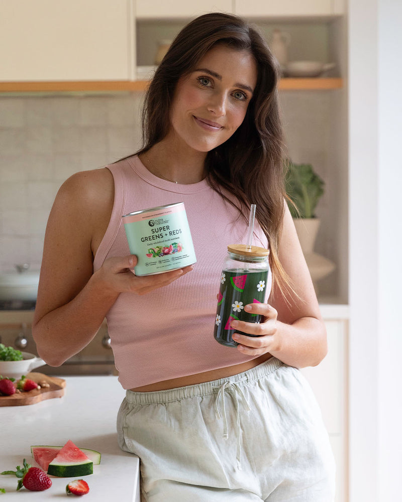 A woman is holding a can of Super Greens + Reds Watermelon Strawberry powder and a dark green glass bottle with Super Greens + Reds Watermelon Strawberry drink