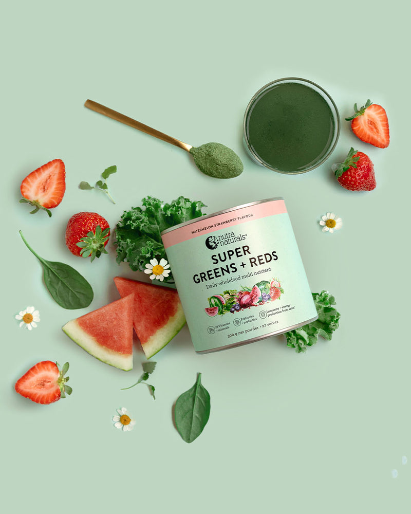 A tin of Super Greens + Reds Watermelon Strawberry powder is styled in a flat lay with fresh strawberries, watermelon slices, kale, spinach, daisies, and a spoonful of green powder on a light sage green background.