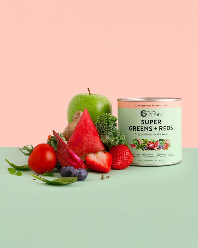A tin of Super Greens + Reds Watermelon Strawberry powder stands on a mint green surface against a peach-pink background, surrounded by fresh fruits and vegetables including a green apple, watermelon slice, tomato, strawberries, blueberries, and broccoli.