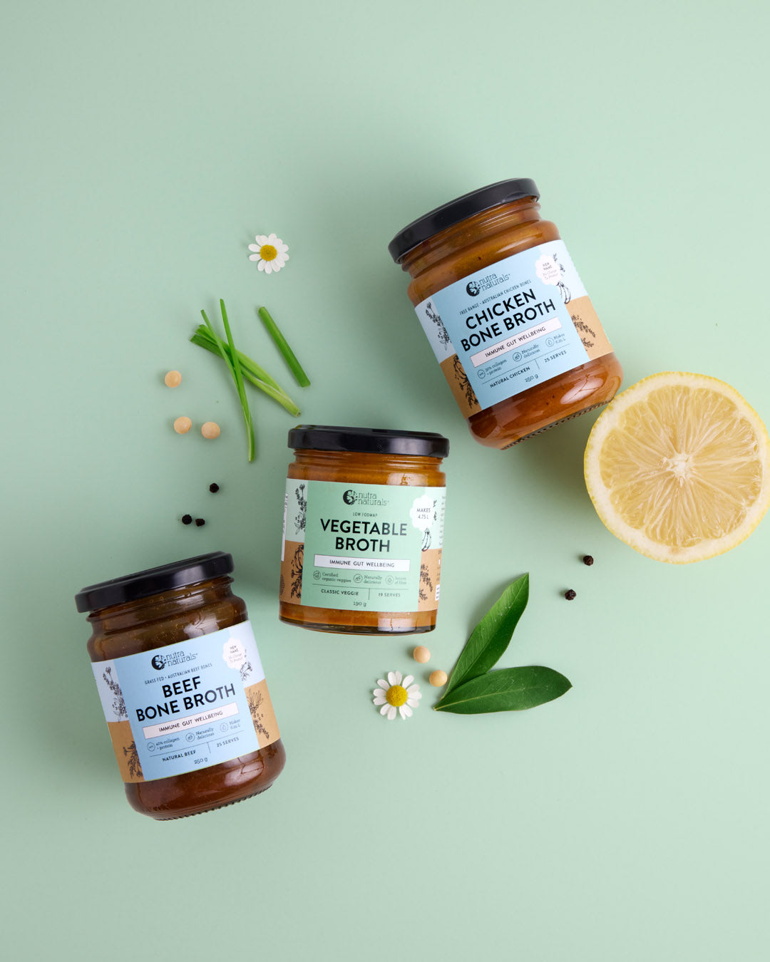 Three jars of bone broth on a light green background with herbs and a lemon slice.